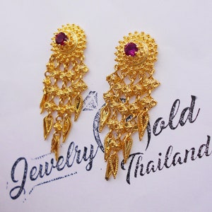 May include: A pair of gold dangle earrings with a pink gem in the center of each earring. The earrings have a delicate, intricate design with multiple layers of small, gold beads and leaves. The earrings are on a white background with the text "Jewelry Gold Thailand" printed in black.