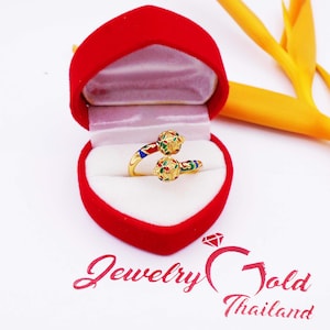May include: A gold ring with two colorful enamel balls. The ring is in a red velvet ring box. The box is on a white surface.