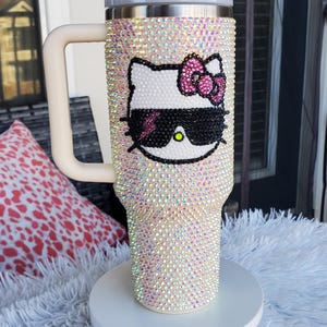 Puede incluir: Un vaso adornado con pedrer&iacute;a con la cara de un gato de dibujos animados con gafas de sol y un lazo rosa. El vaso est&aacute; cubierto de pedrer&iacute;a iridiscente, tiene un asa y una tapa de color crema y se exhibe sobre un soporte blanco.