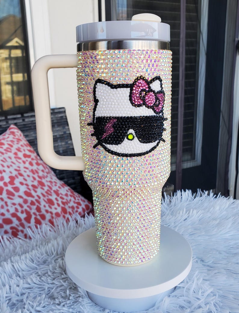 Puede incluir: Un vaso adornado con pedrer&iacute;a con la cara de un gato de dibujos animados con gafas de sol y un lazo rosa. El vaso est&aacute; cubierto de pedrer&iacute;a iridiscente, tiene un asa y una tapa de color crema y se exhibe sobre un soporte blanco.