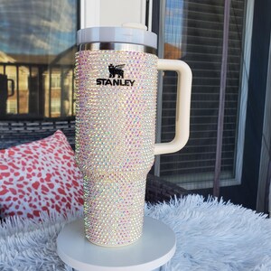 Puede incluir: Un vaso Stanley adornado con pedrer&iacute;a iridiscente, con asa y tapa blancas. El logotipo negro de Stanley es visible. El vaso se exhibe sobre un pedestal blanco. Las medidas no son visibles.