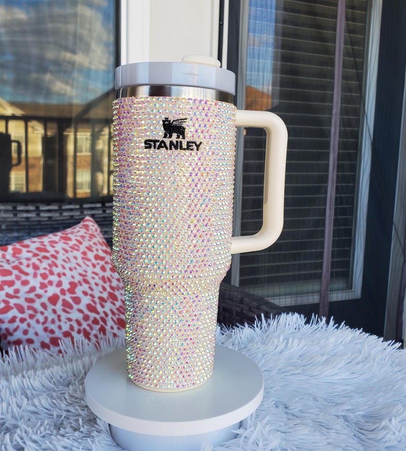 Puede incluir: Un vaso Stanley adornado con pedrer&iacute;a iridiscente, con asa y tapa blancas. El logotipo negro de Stanley es visible. El vaso se exhibe sobre un pedestal blanco. Las medidas no son visibles.