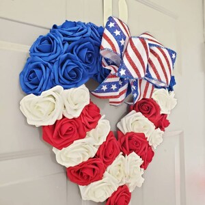 May include: A heart-shaped wreath made of blue, red, and white roses, with a patriotic ribbon bow. The wreath is designed with alternating stripes of red and white roses, and a section of blue roses.