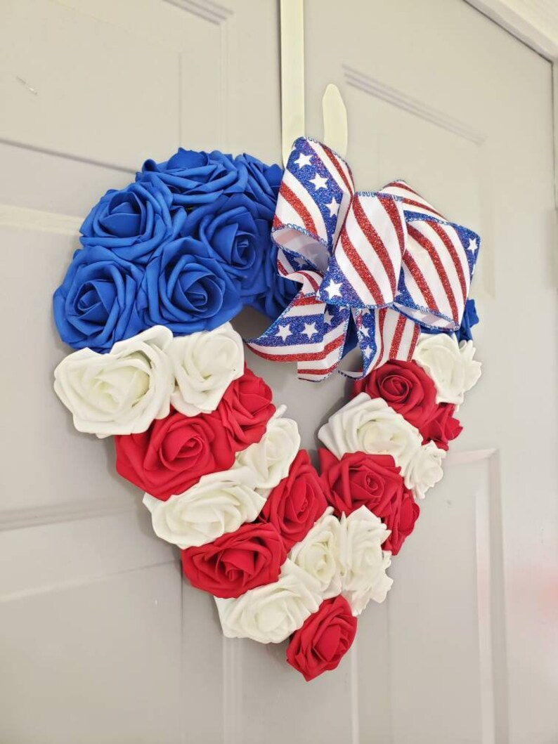 May include: A heart-shaped wreath made of blue, red, and white roses, with a patriotic ribbon bow. The wreath is designed with alternating stripes of red and white roses, and a section of blue roses.