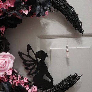 May include: A black crescent moon wreath decorated with pink and black flowers, a black fairy silhouette, and a crystal hanging from a silver string. The wreath has the text "Jess Made Design" at the bottom.