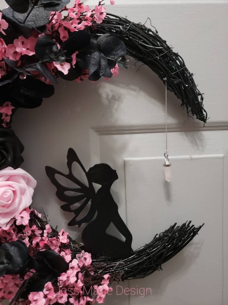 May include: A black crescent moon wreath decorated with pink and black flowers, a black fairy silhouette, and a crystal hanging from a silver string. The wreath has the text "Jess Made Design" at the bottom.