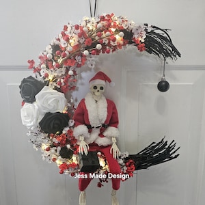 May include: A black and white crescent moon wreath decorated with red and white berries, white lights, and black roses. A skeleton dressed as Santa Claus sits on the wreath holding a black gift box. The wreath has a black ribbon for hanging and a black ornament hanging from the bottom.