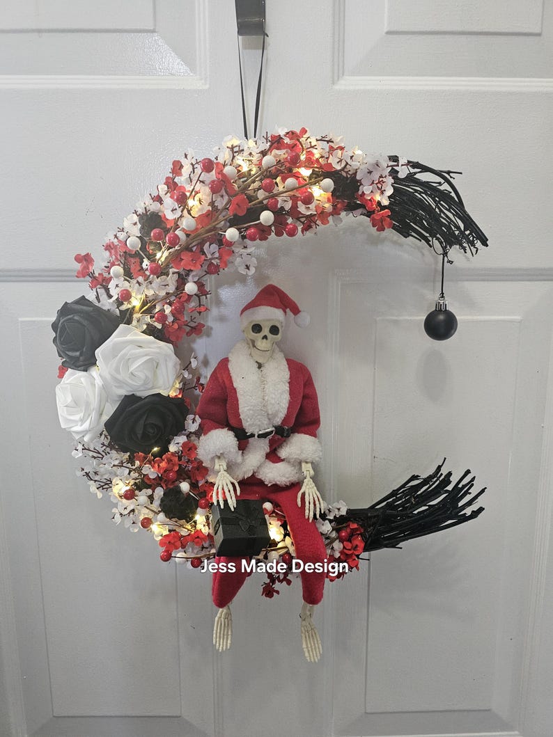 May include: A black and white crescent moon wreath decorated with red and white berries, white lights, and black roses. A skeleton dressed as Santa Claus sits on the wreath holding a black gift box. The wreath has a black ribbon for hanging and a black ornament hanging from the bottom.