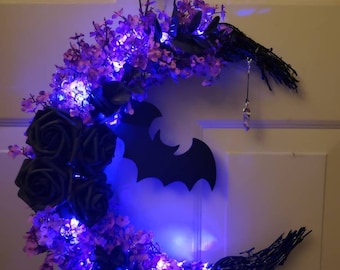 Puede incluir: Una corona de Halloween en forma de media luna de color morado y negro con un recorte de murciélago, rosas negras y luces moradas. La corona está colgada en una puerta blanca. El texto "Jess Made Design" está en la parte inferior de la imagen.