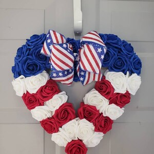 May include: A heart-shaped wreath made of blue, red, and white roses, with a patriotic ribbon bow. The wreath is designed with blue roses at the top, red and white roses forming the heart's shape, and a bow with red stripes and white stars.