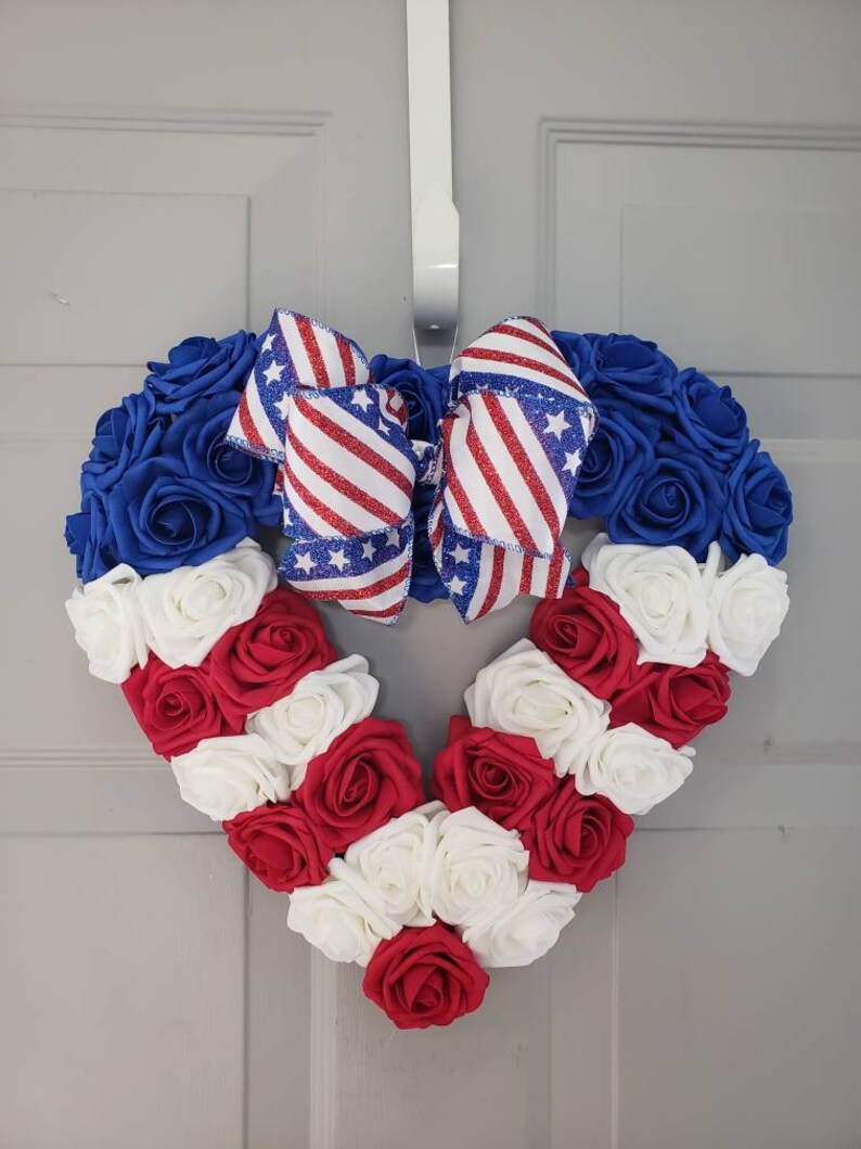 May include: A heart-shaped wreath made of blue, red, and white roses, with a patriotic ribbon bow. The wreath is designed with blue roses at the top, red and white roses forming the heart's shape, and a bow with red stripes and white stars.