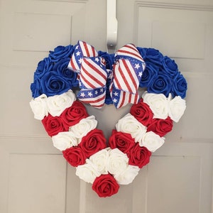 May include: A heart-shaped wreath made of blue, red, and white roses, with a patriotic bow. The wreath is decorated with red and white striped ribbon and blue ribbon with white stars. The wreath is hanging on a white door.