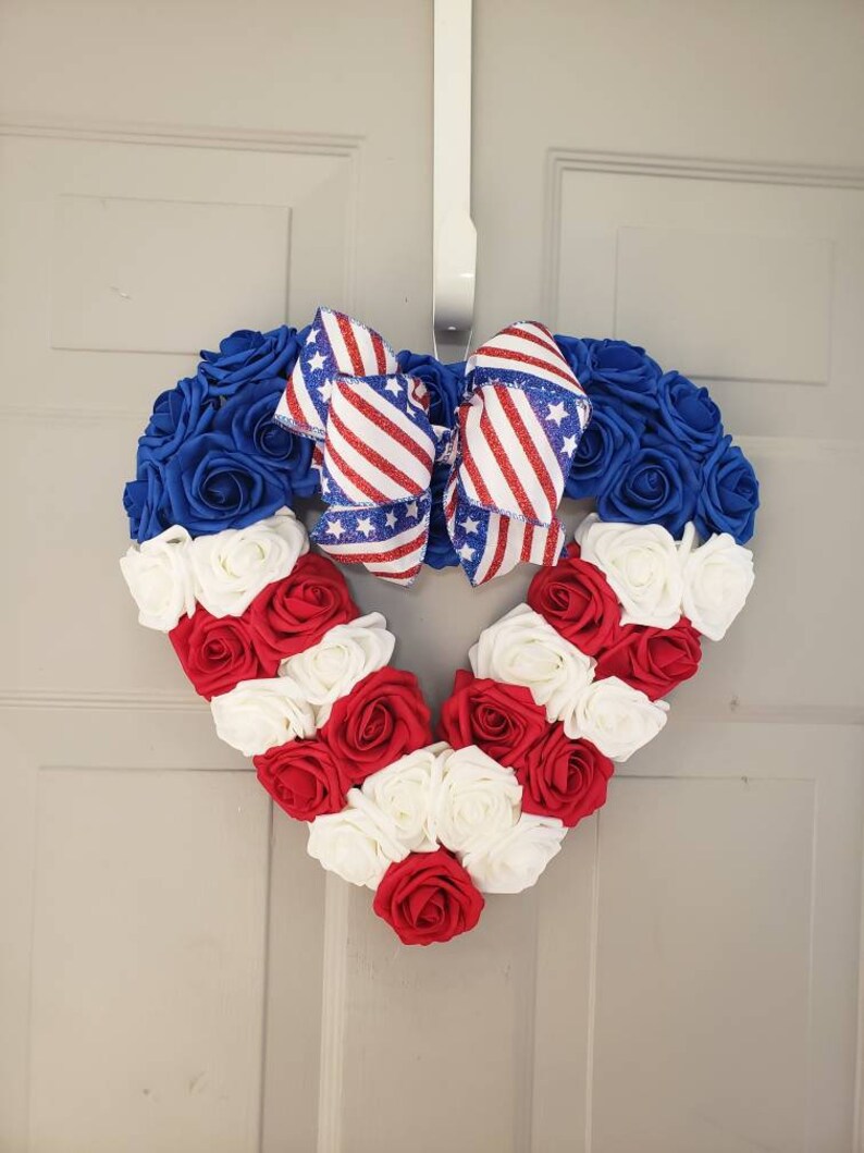 May include: A heart-shaped wreath made of blue, red, and white roses, with a patriotic bow. The wreath is decorated with red and white striped ribbon and blue ribbon with white stars. The wreath is hanging on a white door.