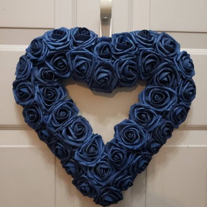 K&ouml;nnte beinhalten: Ein herzf&ouml;rmiger Kranz aus blauen Seidenrosen. Der Kranz h&auml;ngt an einer wei&szlig;en T&uuml;r.