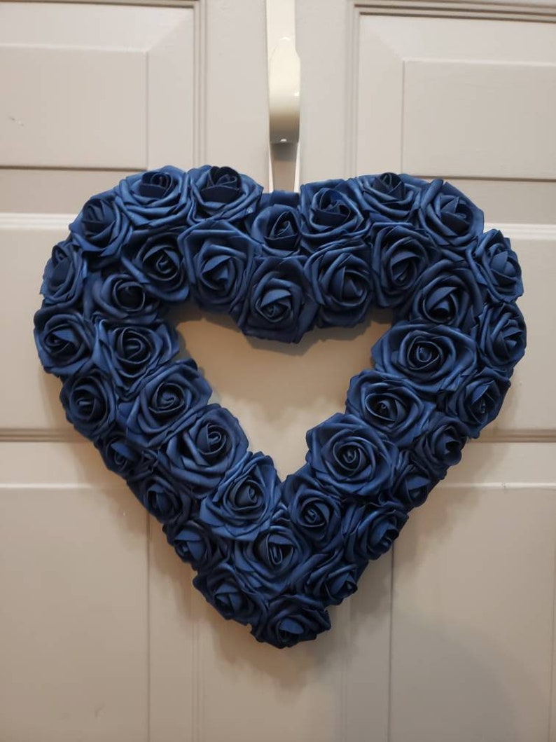 K&ouml;nnte beinhalten: Ein herzf&ouml;rmiger Kranz aus blauen Seidenrosen. Der Kranz h&auml;ngt an einer wei&szlig;en T&uuml;r.