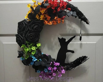 Puede incluir: Una silueta de gato negro posada en una corona en forma de media luna decorada con rosas negras, flores de colores arcoíris y un cristal negro colgando de un cordón negro. La corona está hecha de ramitas y tiene el texto "Jess Made Design" en la parte inferior.