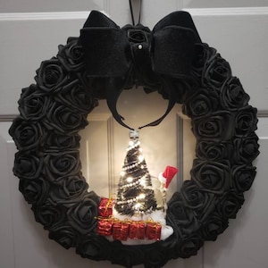 May include: A black wreath made of fabric roses with a black velvet bow. A small, lit Christmas tree with red presents sits in the center of the wreath.
