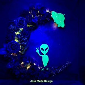 May include: A black and blue floral wreath with a glowing green alien and UFO. The wreath is shaped like a crescent moon. The text "Jess Made Design" is at the bottom of the image.
