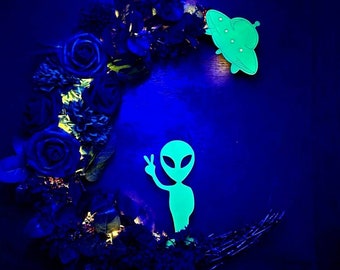 Puede incluir: Una corona floral negra y azul con un extraterrestre y un OVNI verde brillante. La corona tiene forma de media luna. El texto "Jess Made Design" está en la parte inferior de la imagen.