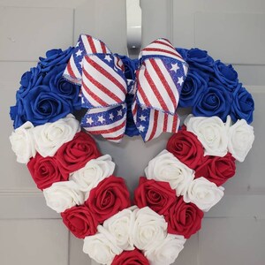 May include: A heart-shaped wreath composed of blue, red, and white roses. A large bow with red and white stripes and blue stars is at the top. The wreath is hanging on a white door.