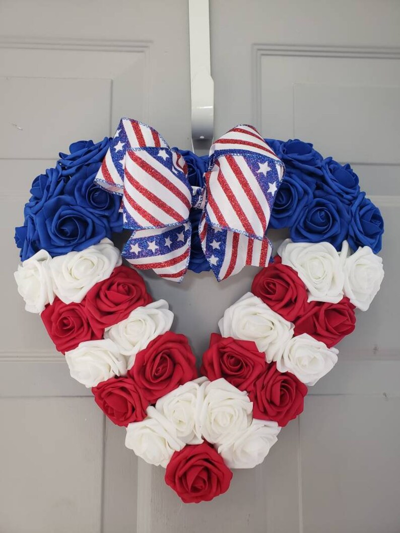 May include: A heart-shaped wreath composed of blue, red, and white roses. A large bow with red and white stripes and blue stars is at the top. The wreath is hanging on a white door.