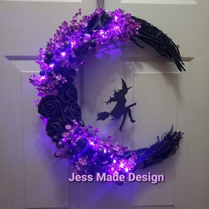 May include: A black crescent moon wreath decorated with purple flowers and black roses. The wreath has purple fairy lights and a black silhouette of a witch flying on a broomstick. The wreath has the text "Jess Made Design" at the bottom.