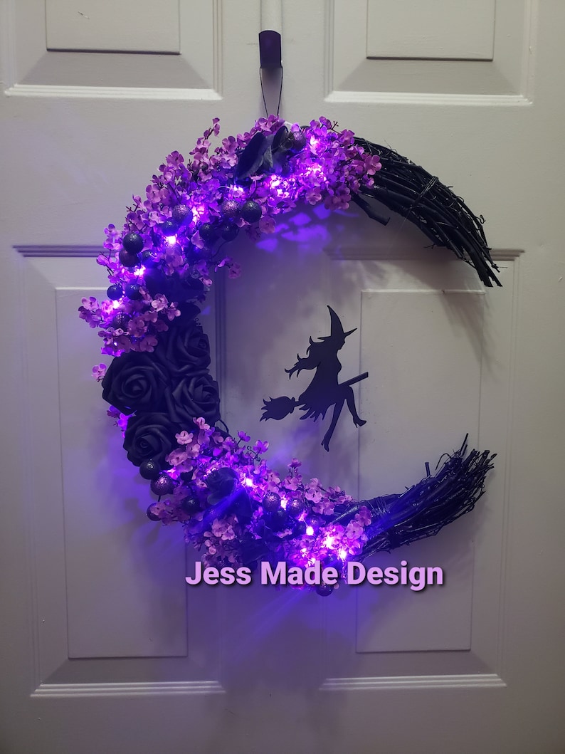 May include: A black crescent moon wreath decorated with purple flowers and black roses. The wreath has purple fairy lights and a black silhouette of a witch flying on a broomstick. The wreath has the text "Jess Made Design" at the bottom.