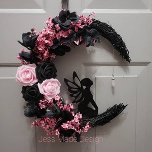 May include: A black crescent moon wreath decorated with pink and black flowers, a black fairy silhouette, and a crystal hanging from the bottom. The wreath is labeled "Jess Made Design".