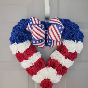 May include: A heart-shaped wreath made of blue, red, and white roses, with a patriotic bow. The wreath is designed with blue roses around the top, red and white roses forming the bottom, and a bow with red and white stripes and blue stars.