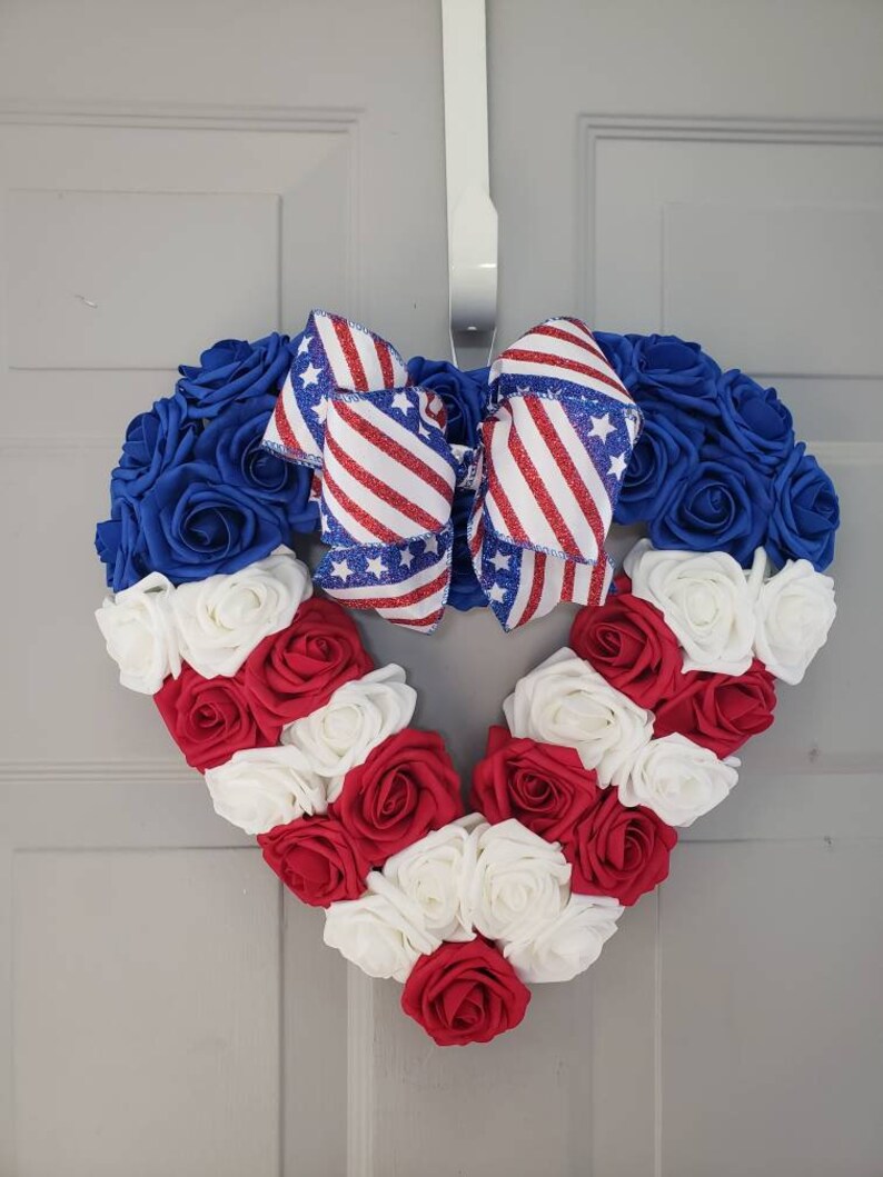 May include: A heart-shaped wreath made of blue, red, and white roses, with a patriotic bow. The wreath is designed with blue roses around the top, red and white roses forming the bottom, and a bow with red and white stripes and blue stars.