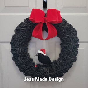 May include: A black rose wreath with a red velvet bow and a black crow wearing a Santa hat. The wreath is hanging on a white door. The text "Jess Made Design" is visible at the bottom of the image.