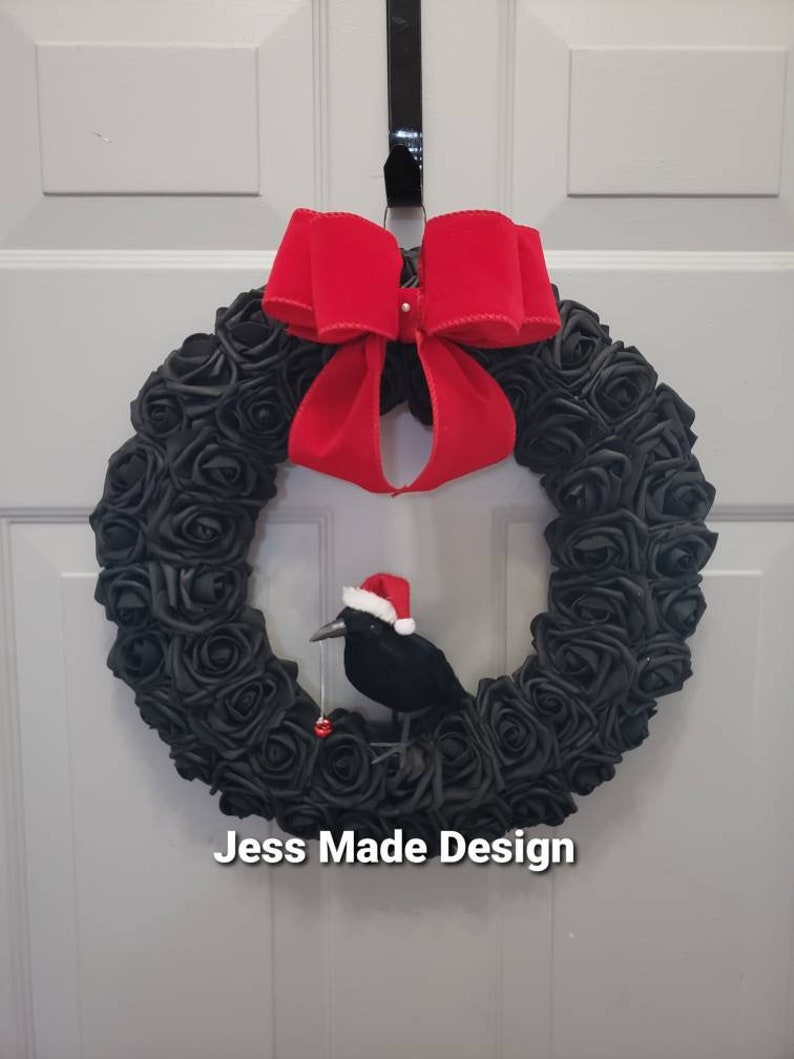 May include: A black rose wreath with a red velvet bow and a black crow wearing a Santa hat. The wreath is hanging on a white door. The text "Jess Made Design" is visible at the bottom of the image.