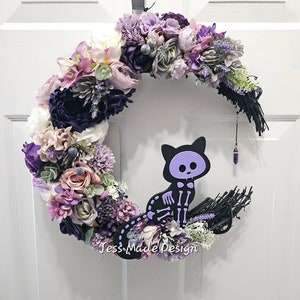 Puede incluir: Una corona floral de color morado y blanco con forma de media luna con una silueta de gato esqueleto negro en el centro. La corona est&aacute; decorada con un colgante de cristal morado y el texto "Jess Made Design".