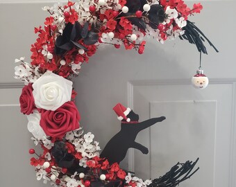 Puede incluir: Una corona de Navidad roja y blanca con forma de media luna con una silueta de gato negro que lleva un gorro de Papá Noel. La corona está decorada con flores rojas y blancas, bayas y hojas negras. El texto "Jess Made Design" es visible en la parte inferior de la imagen.