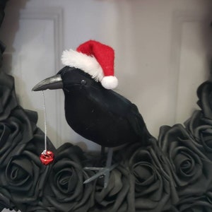 May include: A black crow wearing a red and white Santa hat sits on a wreath of black roses. A red bell hangs from the crow's beak. The wreath has the text "Jess Made Design" at the bottom.