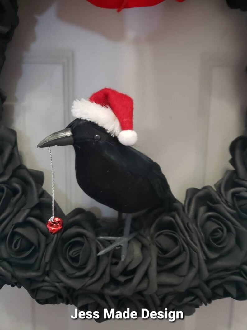 May include: A black crow wearing a red and white Santa hat sits on a wreath of black roses. A red bell hangs from the crow's beak. The wreath has the text "Jess Made Design" at the bottom.