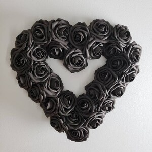 K&ouml;nnte beinhalten: Ein herzf&ouml;rmiger Kranz aus dunkelgrauen Rosen. Die Rosen sind dicht gepackt und bilden die Form eines Herzens mit einer offenen Mitte. Der Kranz ist an einer wei&szlig;en Wand befestigt, was einen starken Kontrast erzeugt.