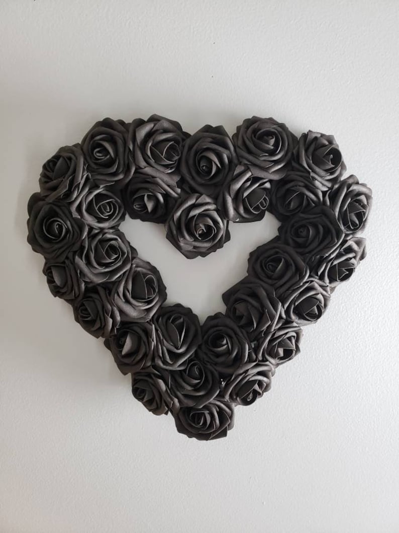 K&ouml;nnte beinhalten: Ein herzf&ouml;rmiger Kranz aus dunkelgrauen Rosen. Die Rosen sind dicht gepackt und bilden die Form eines Herzens mit einer offenen Mitte. Der Kranz ist an einer wei&szlig;en Wand befestigt, was einen starken Kontrast erzeugt.