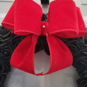 May include: A black wreath with a large red velvet bow. The bow is tied with a white pearl button in the center. The wreath is decorated with black fabric roses. The text "Jess Made Design" is visible at the bottom of the image.
