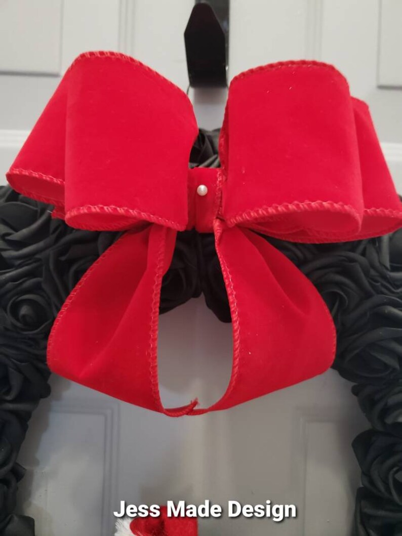 May include: A black wreath with a large red velvet bow. The bow is tied with a white pearl button in the center. The wreath is decorated with black fabric roses. The text "Jess Made Design" is visible at the bottom of the image.