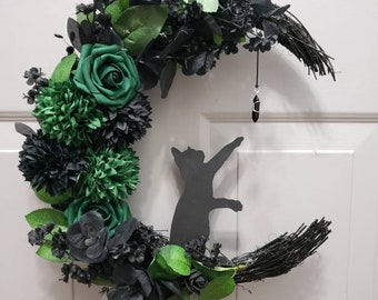 May include: A crescent moon-shaped wreath with a black cat silhouette, adorned with green and black flowers and foliage. A black crystal hangs from the moon's tip. The text "Jess Made Design" is visible at the bottom.