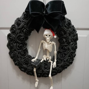 May include: A black rose wreath with a skeleton wearing a Santa hat sitting on it. The skeleton is holding a small gift box.
