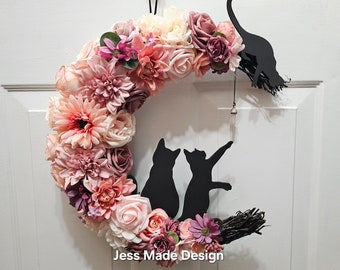 May include: A pink and white floral wreath shaped like a crescent moon with two black cat silhouettes on it. The wreath is decorated with a pink and white floral arrangement and a small bell charm. The text "Jess. Made Design" is at the bottom of the wreath.