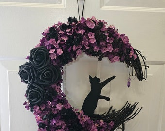 May include: A crescent moon-shaped wreath with a black cat silhouette, black roses, and purple flowers. The wreath is decorated with a small purple crystal and is hanging on a white door. The text "Jess Made Design" is visible.
