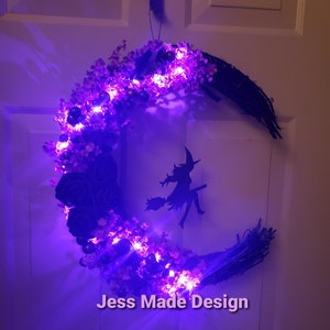 May include: A purple and black crescent moon wreath with a witch silhouette and purple lights. The wreath is decorated with faux flowers and has the text "Jess Made Design" at the bottom.