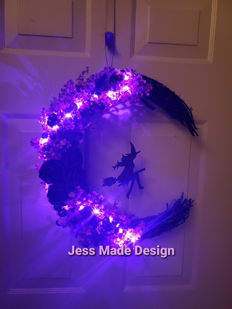 May include: A purple and black crescent moon wreath with a witch silhouette and purple lights. The wreath is decorated with faux flowers and has the text "Jess Made Design" at the bottom.