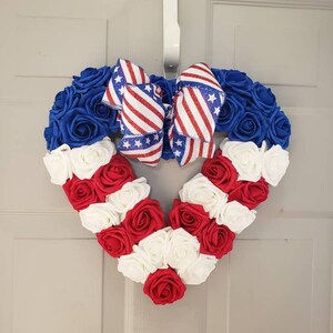 May include: A heart-shaped wreath made of blue, red, and white roses, with a patriotic bow. The wreath is designed with blue roses at the top, red and white roses forming stripes, and a bow with red and white stripes and stars.
