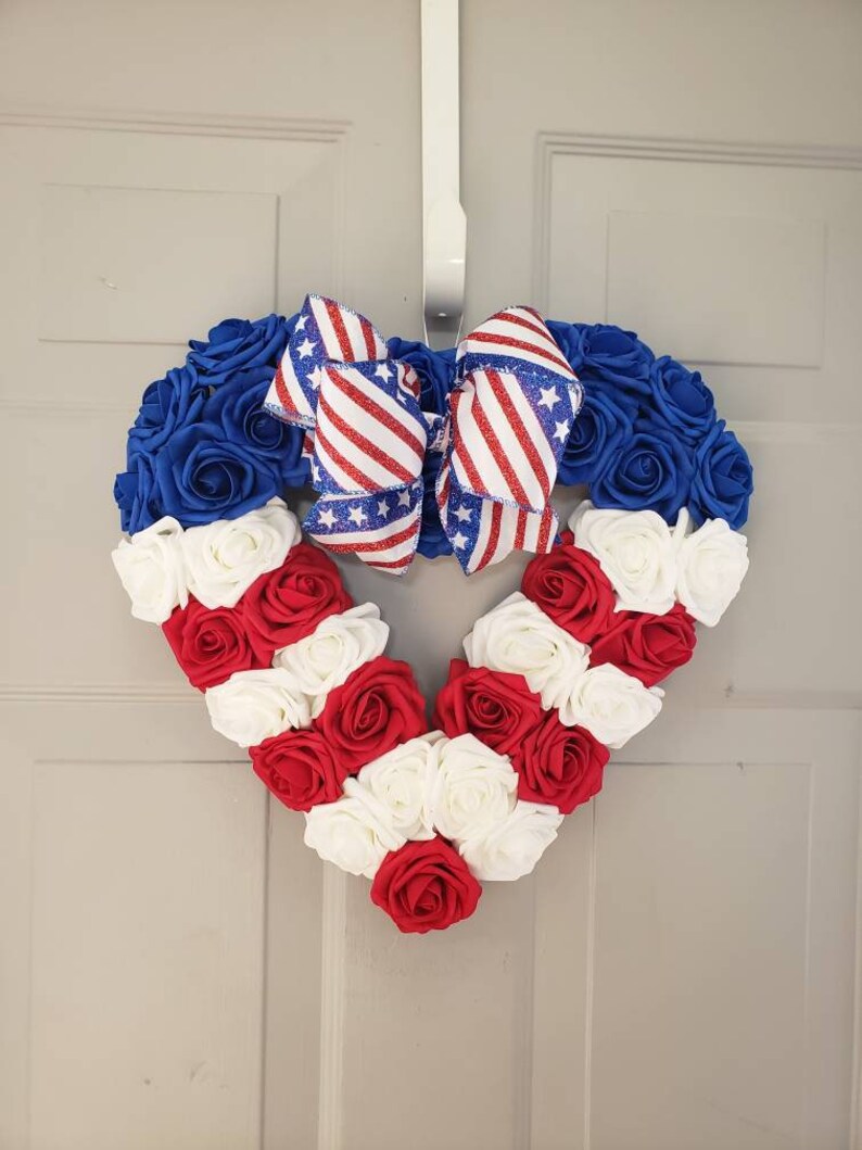 May include: A heart-shaped wreath made of blue, red, and white roses, with a patriotic bow. The wreath is designed with blue roses at the top, red and white roses forming stripes, and a bow with red and white stripes and stars.