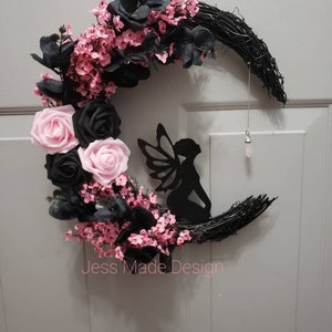 May include: A black crescent moon wreath decorated with pink and black flowers, a black fairy silhouette, and a crystal pendant. The wreath is made of grapevine and has a "Jess Made Design" label.
