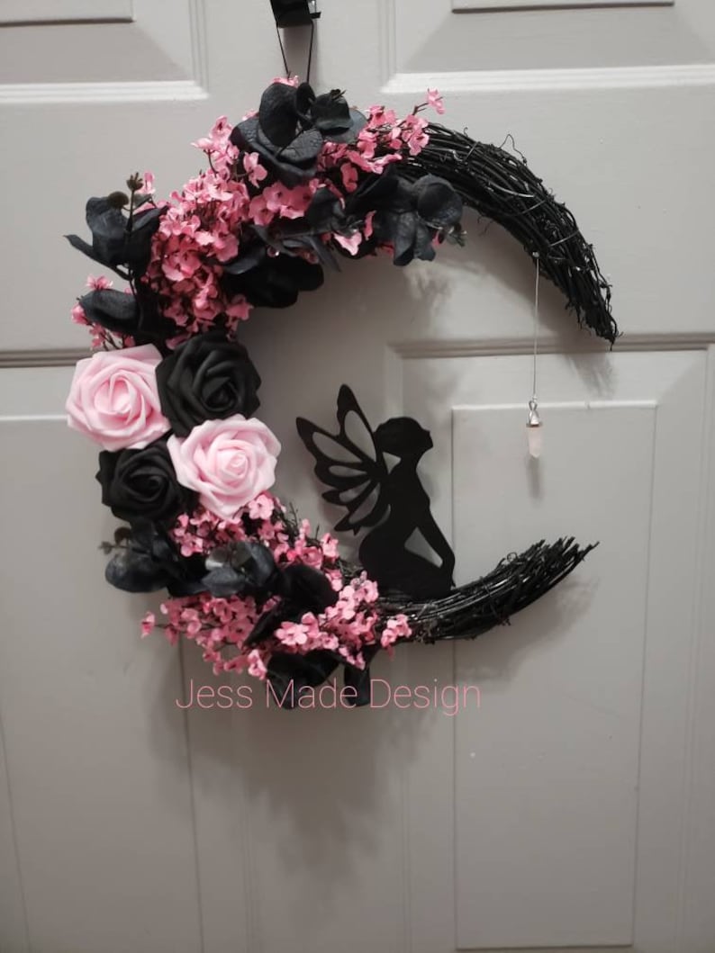 May include: A black crescent moon wreath decorated with pink and black flowers, a black fairy silhouette, and a crystal pendant. The wreath is made of grapevine and has a "Jess Made Design" label.